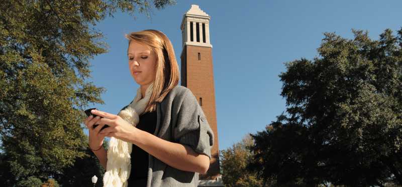 A student looking at their mobile phone while standing in front of Denny Chimes tower at the University of Alabama in Tuscaloosa.
