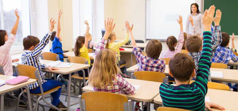 Students raising their hands with teacher at the front of classroom.