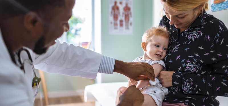 A toddler in a person's lap while another person gives the child a vaccine.