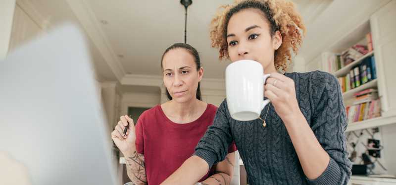Two people in home are looking intently at laptop while one drinks out of coffee mug.