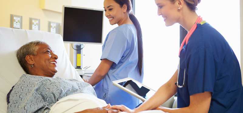Two women in scrubs with older female patient in hospital bed.