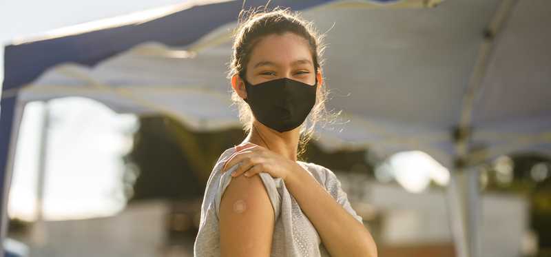 A masked person holds up a sleeve to show vaccination bandage.