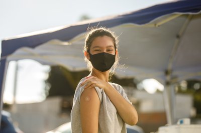 A masked person holds up a sleeve to show vaccination bandage.