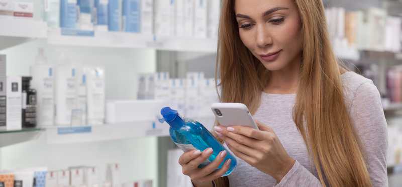 Person standing in pharmacy aisle is holding mouthwash and looking at phone.