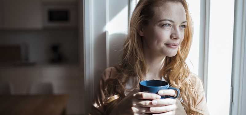 Person holding coffee mug and looking out window with a smile.