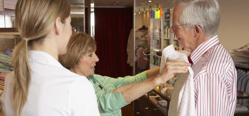 A woman holding shirt against older man's body in store.
