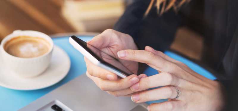 Woman uses her smartphone with laptop in front of her and coffee at her side.
