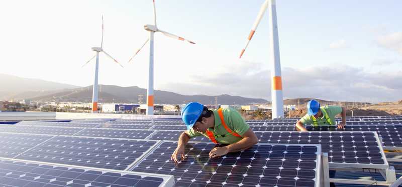 Workers on solar panels with wind turbines in background.