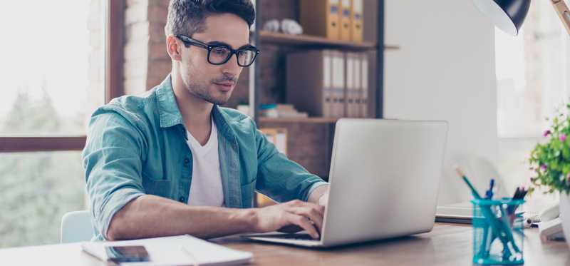 Young male adult in glasses typing on a laptop.