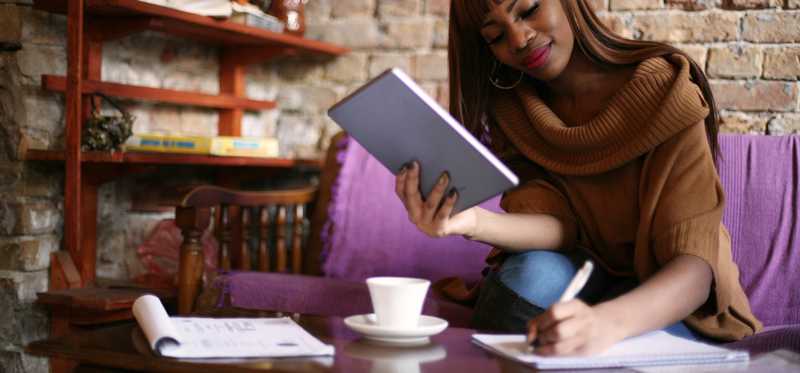Person sitting at coffee table holding tablet and writing on notepad.