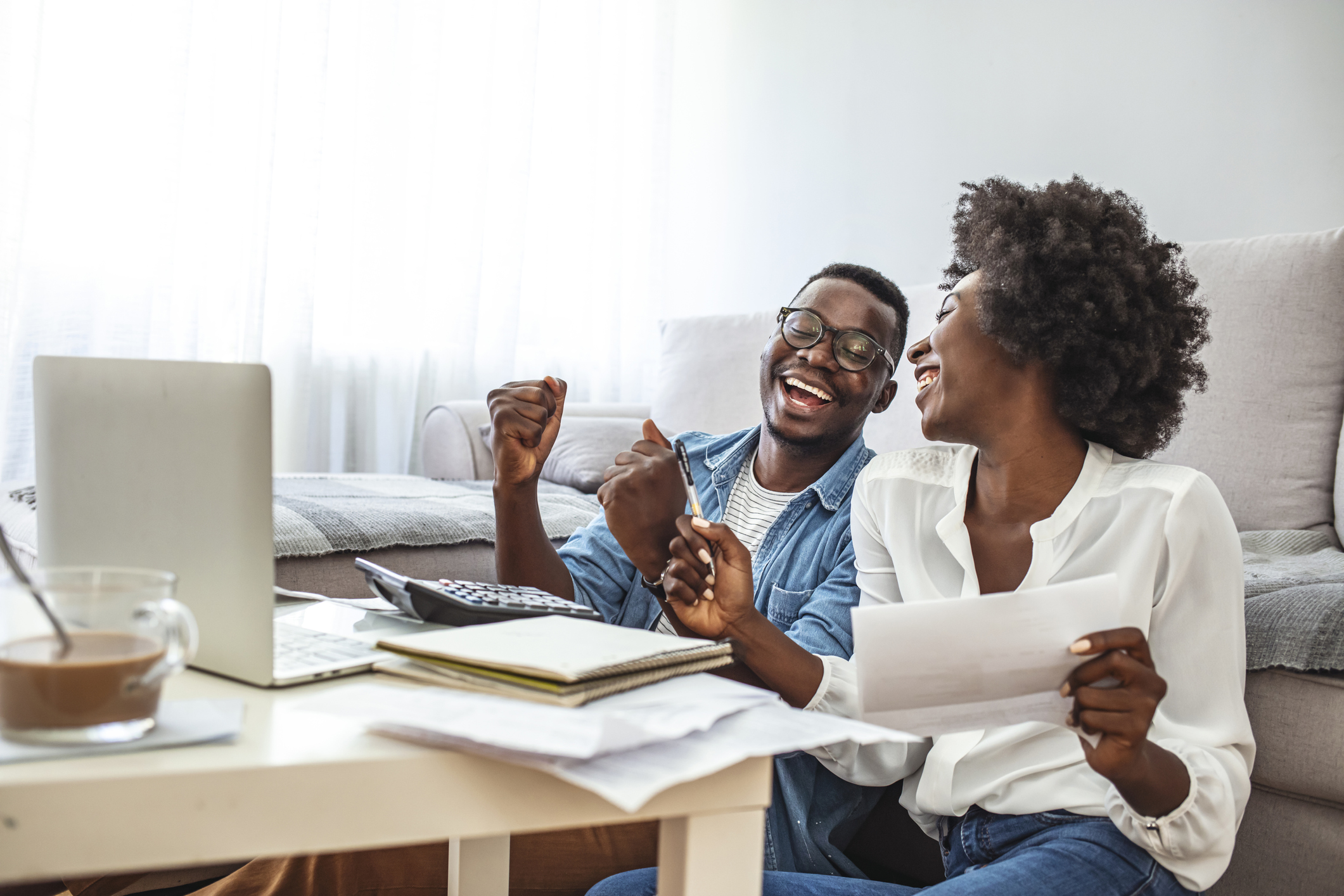 13-couple-personal-finance Two adults sitting on floor in front of coffee table are celebrating.