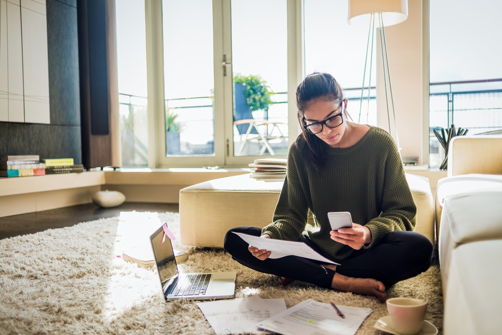2-younger-woman-budgets.jpg Person wearing glasses sits on floor and reviews paperwork thoughtfully.