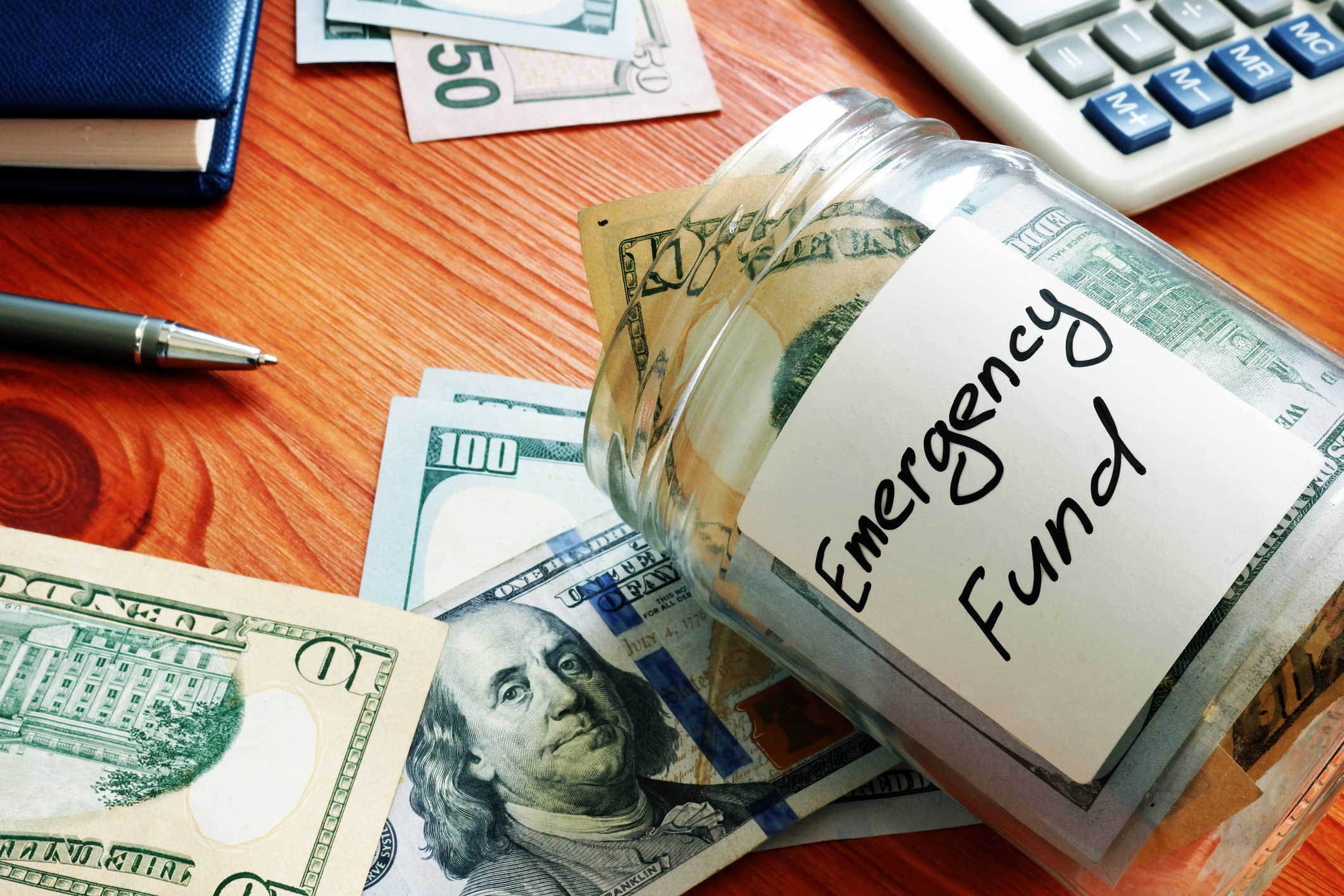 Emergency Fund A desk with a calculator and a glass jar that says Emergency Fund and is full of cash.