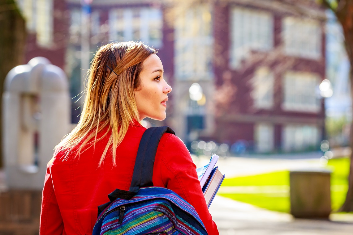 Young person with backpack and books walking toward school building.
