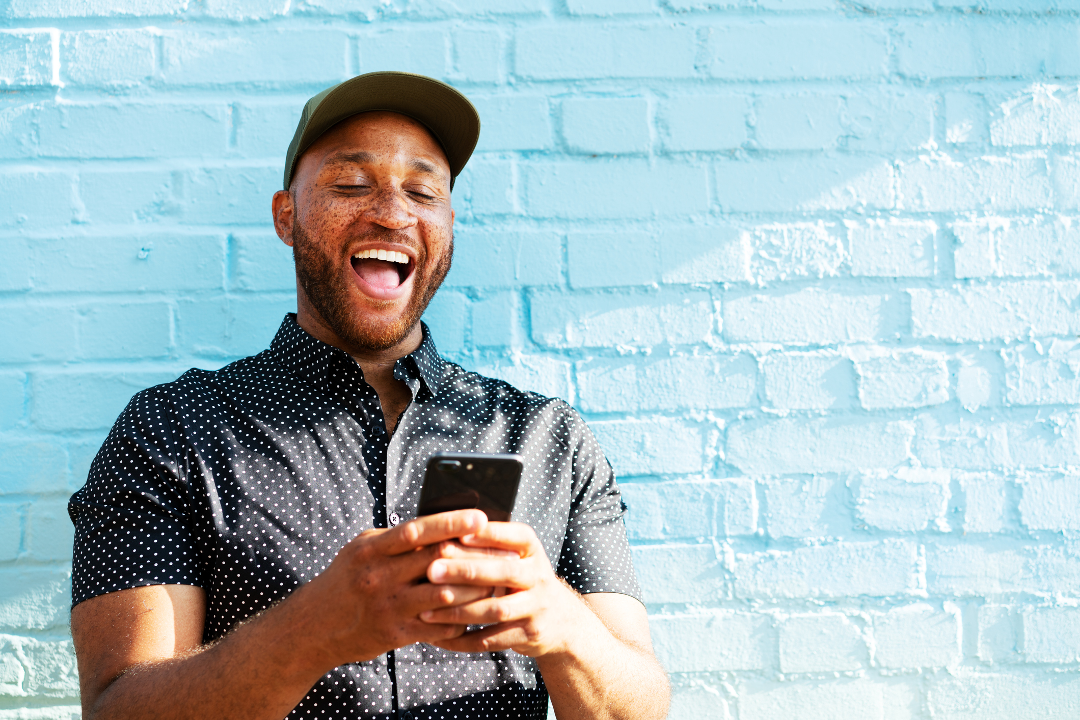 8-happy-adult-smartphone Individual wearing baseball cap smiles broadly while looking at smartphone screen outside.
