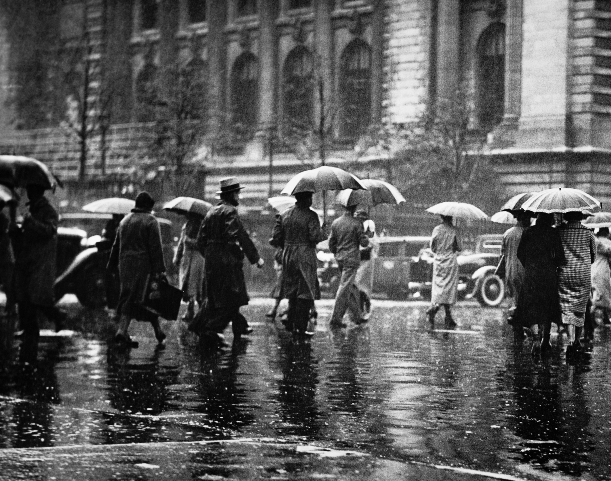 In black and white, several people walk across a parking lot in the rain holding umbrellas in older style clothing.