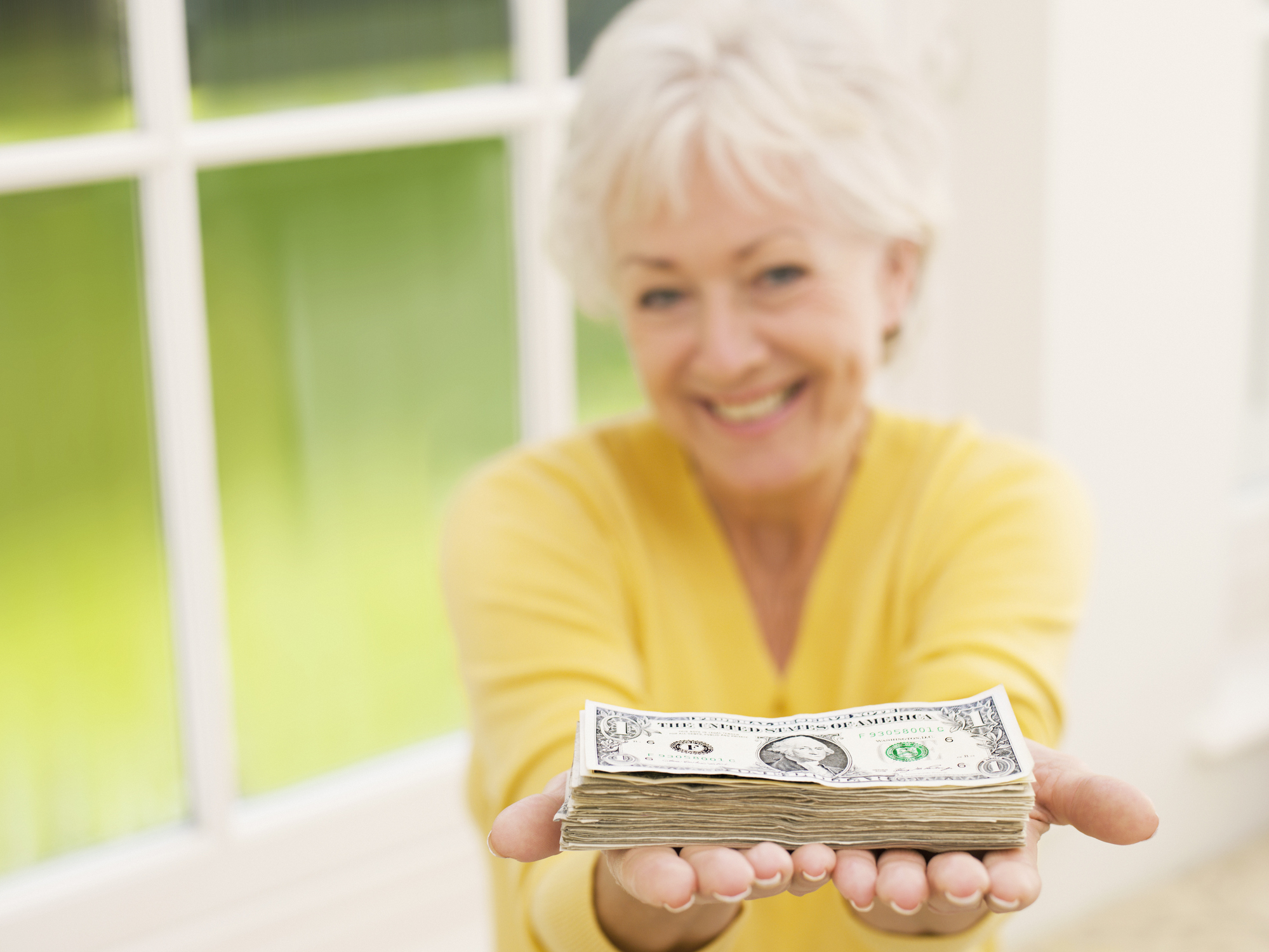 A happy senior woman holding a neat stack of cash bills in her outstretched hands.