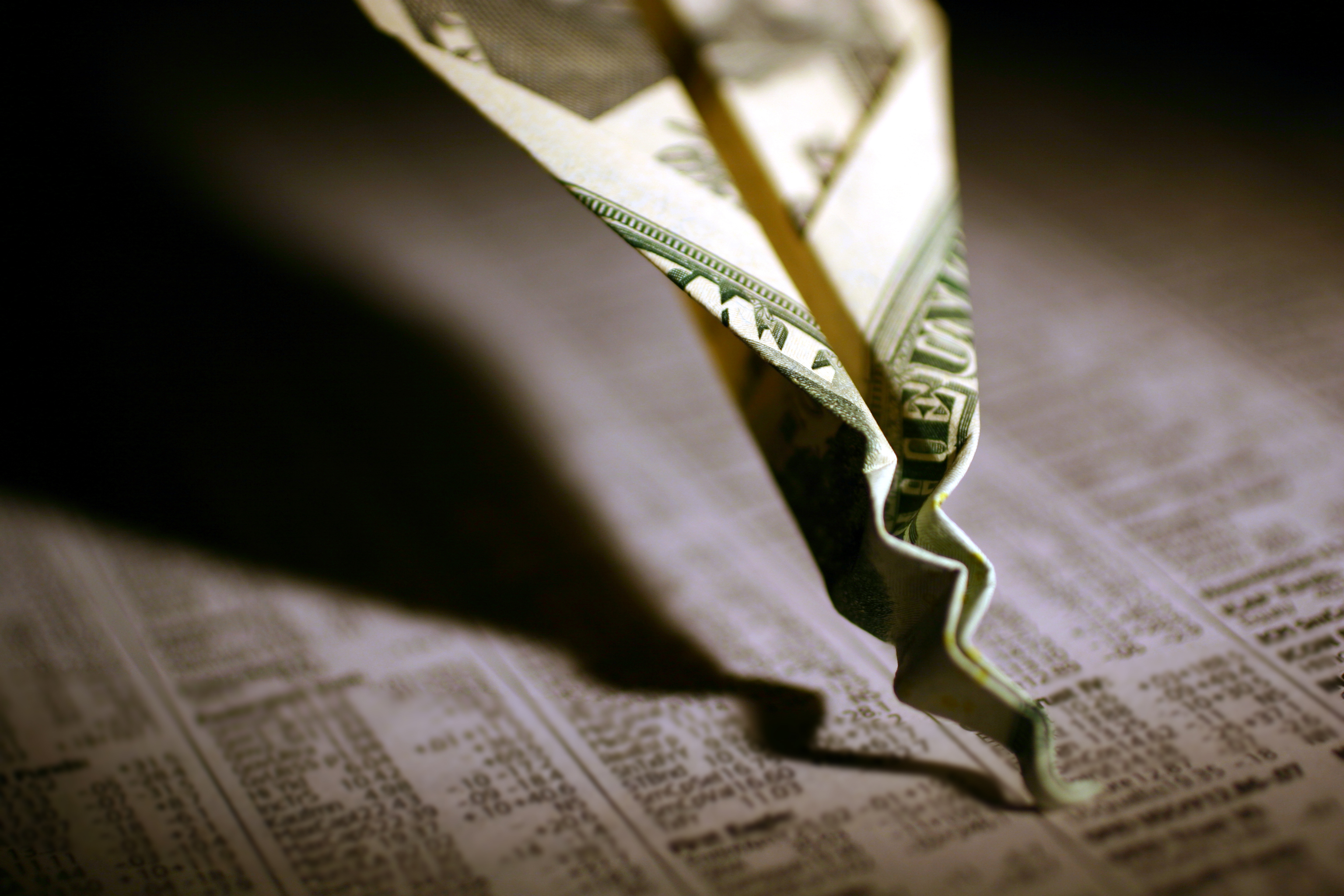 A paper airplane made out of a one dollar bill that's crashed and crumpled into the financial section of the newspaper.