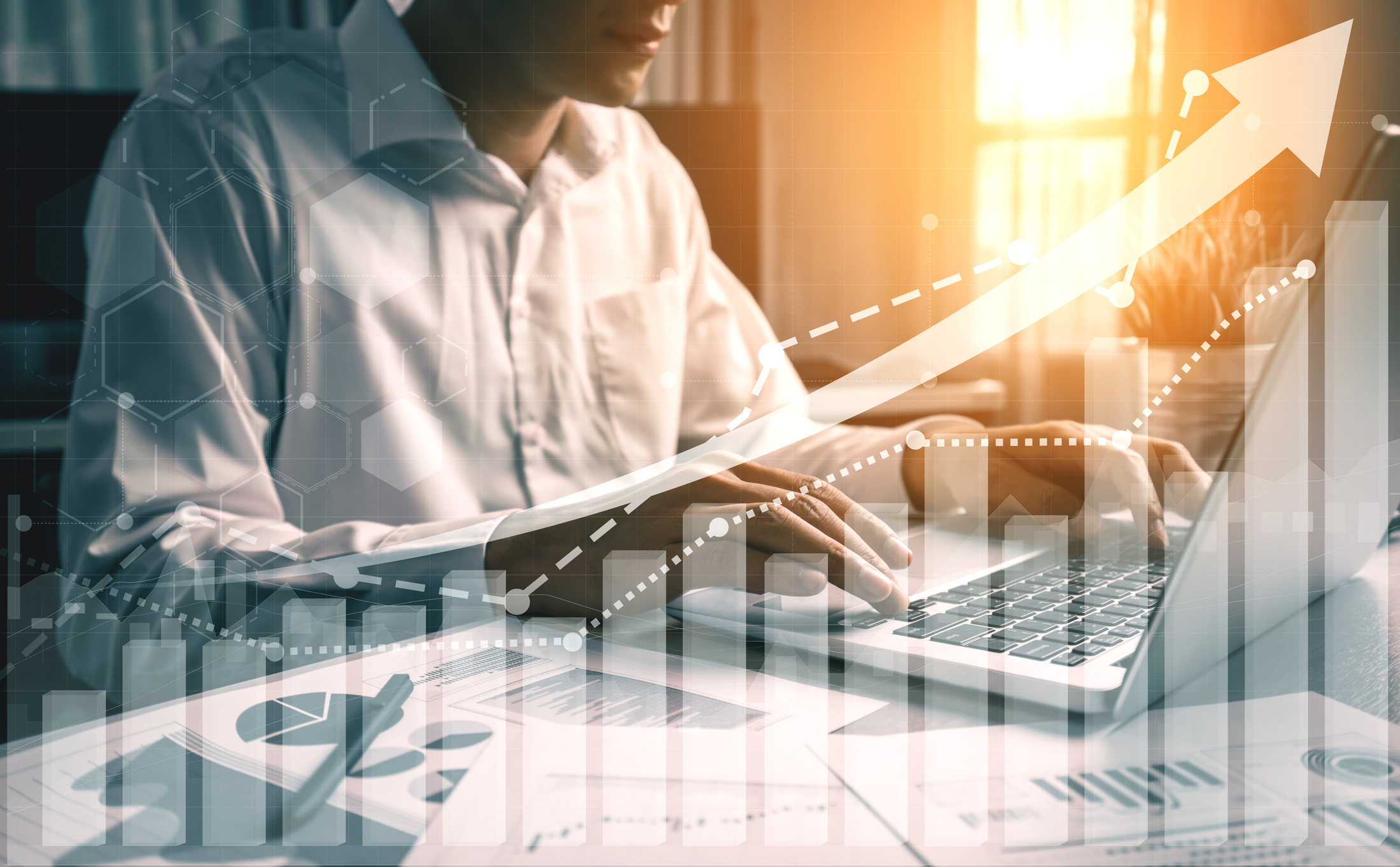 A person at a computer with financial charts on a desk overlaid by an upward pointing arrow.