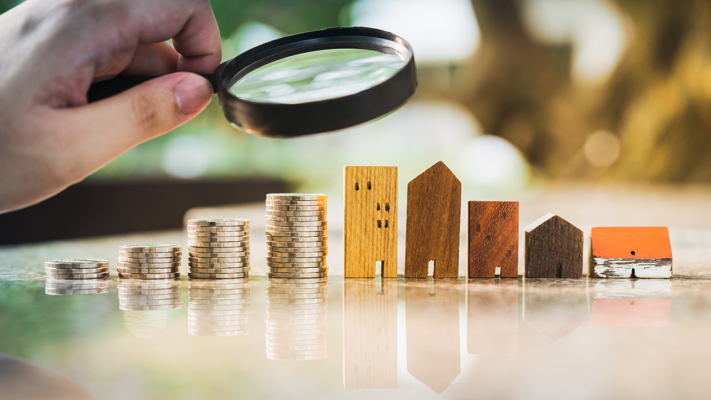 A person holding a magnifying glass looking at a row of rising coins and buildings. A person holding a magnifying glass looking at a row of rising coins and buildings.