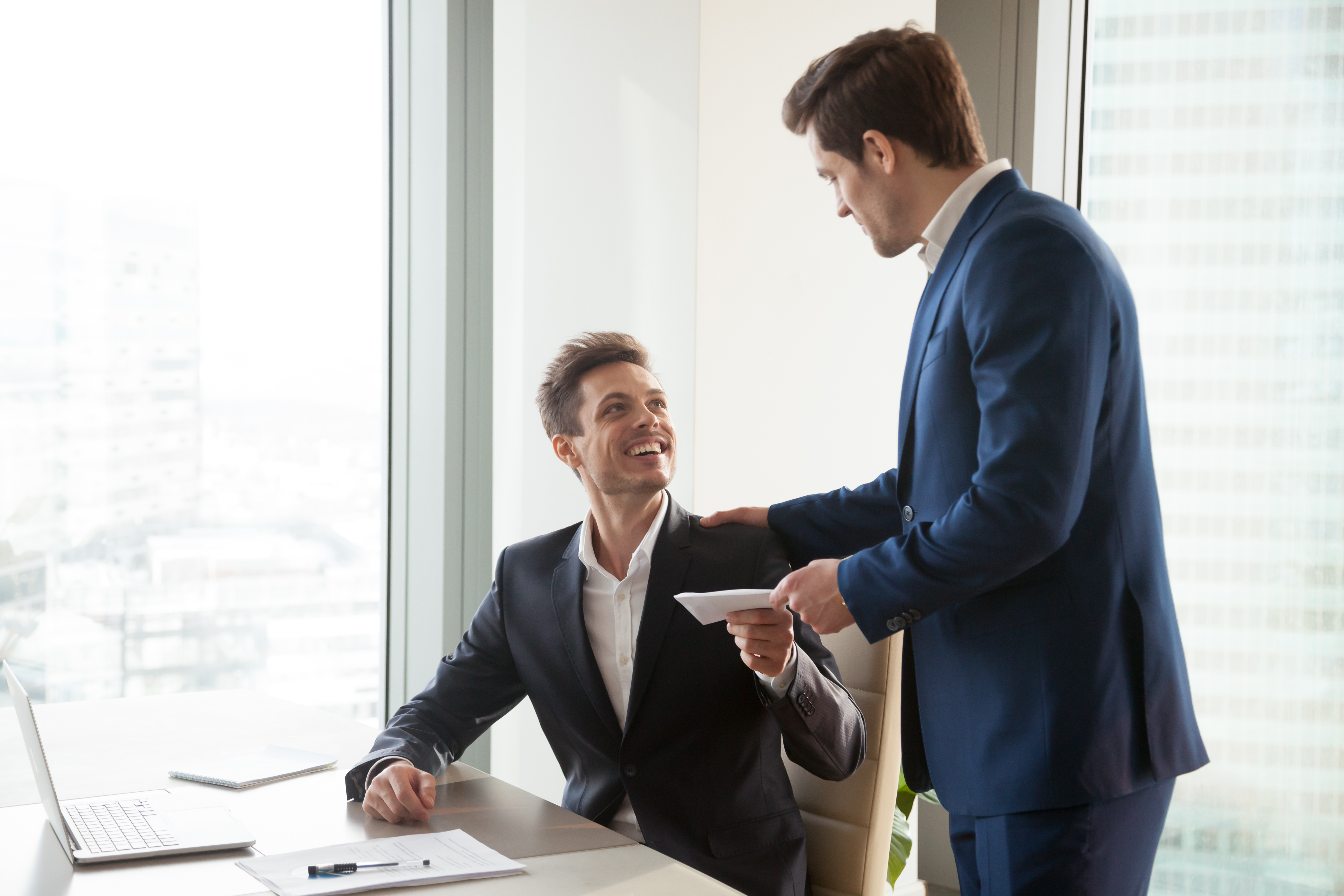 Boss handing envelope of money to employee A standing person hands an envelope to a smiling person seated at a desk.