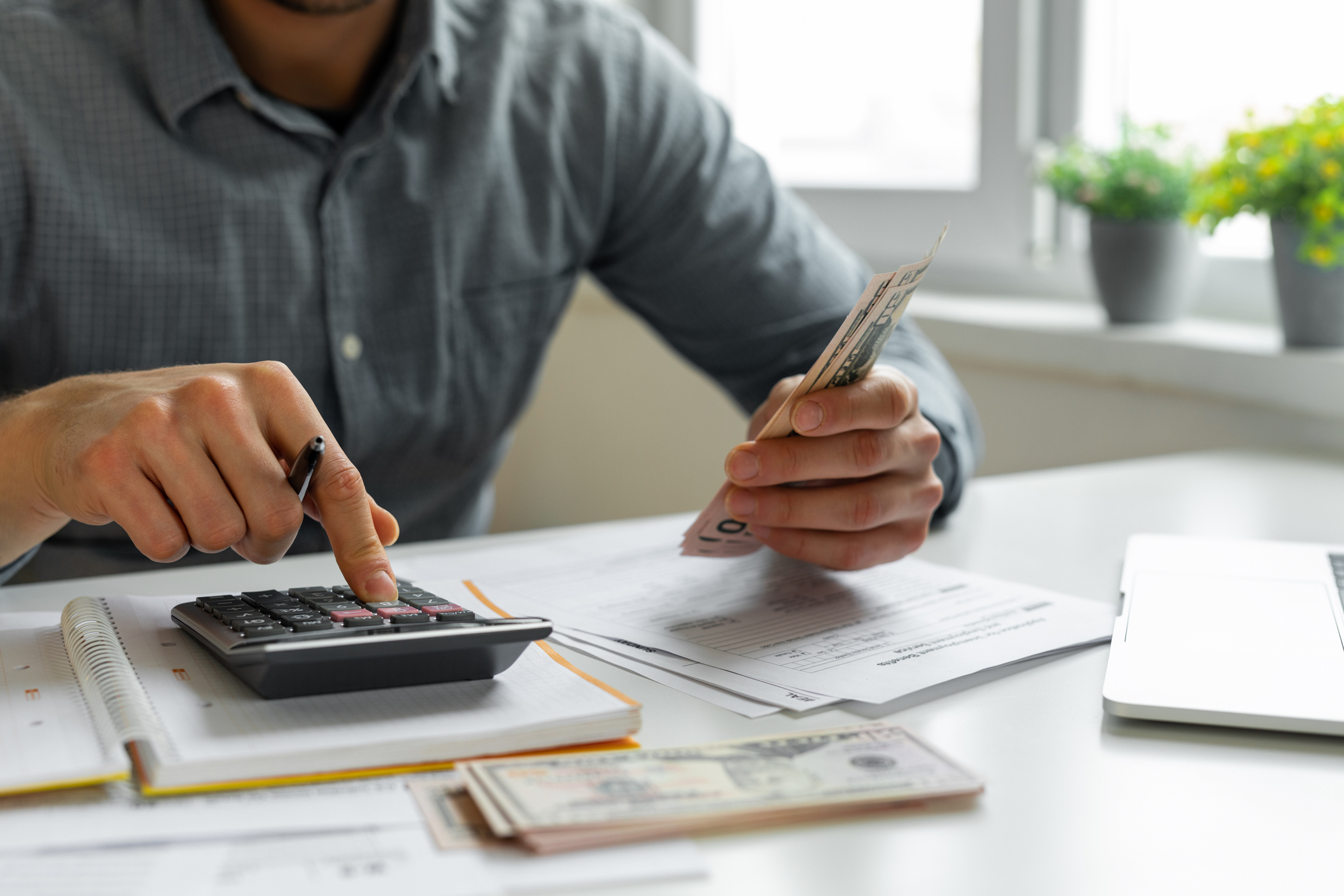 GettyImages-1309118129.jpg Person using calculator while holding cash and looking at papers on a desk.