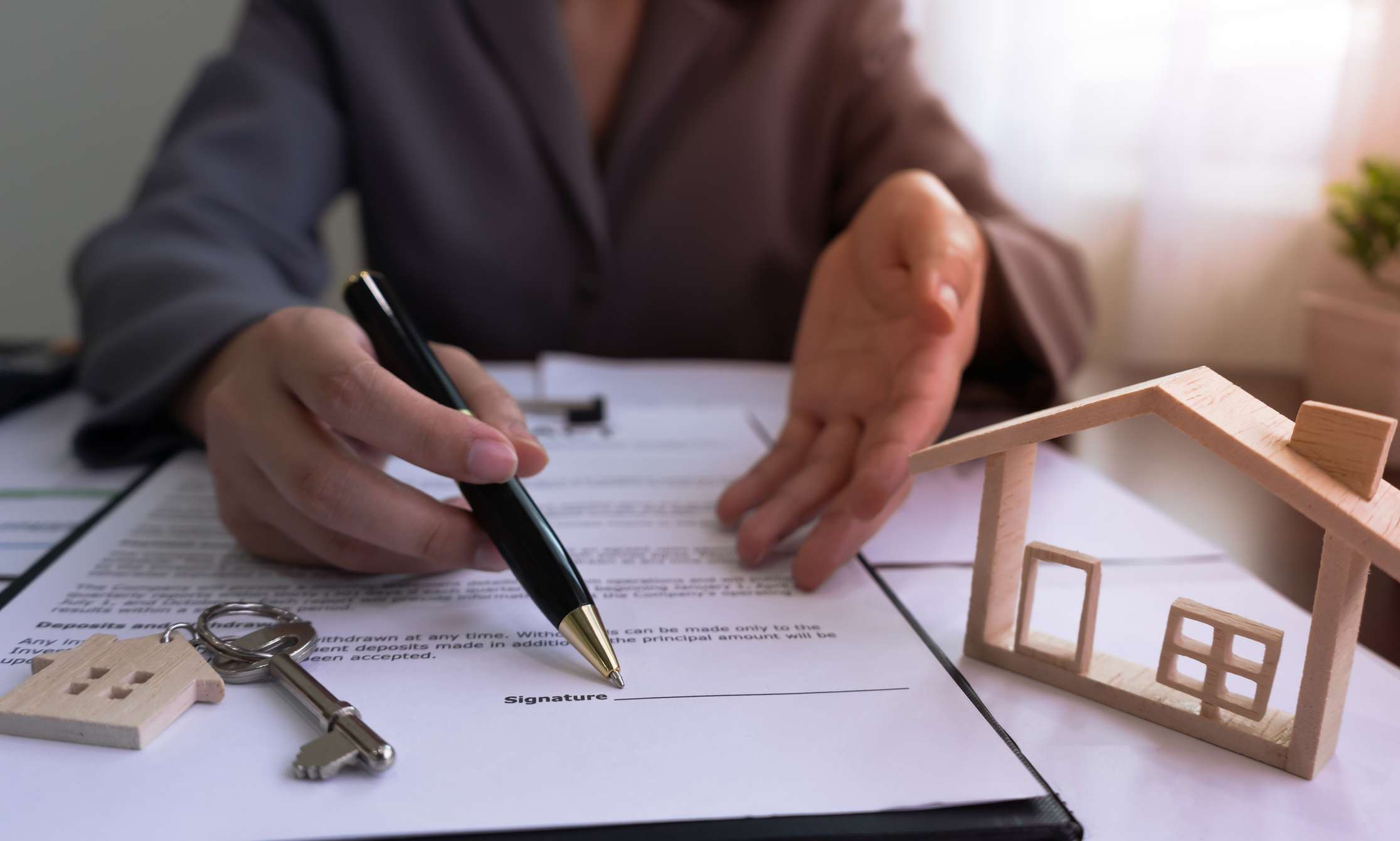 Person is sitting across a desk and using a pen to show you where to sign on a document that is sitting next to a house key and miniature home.