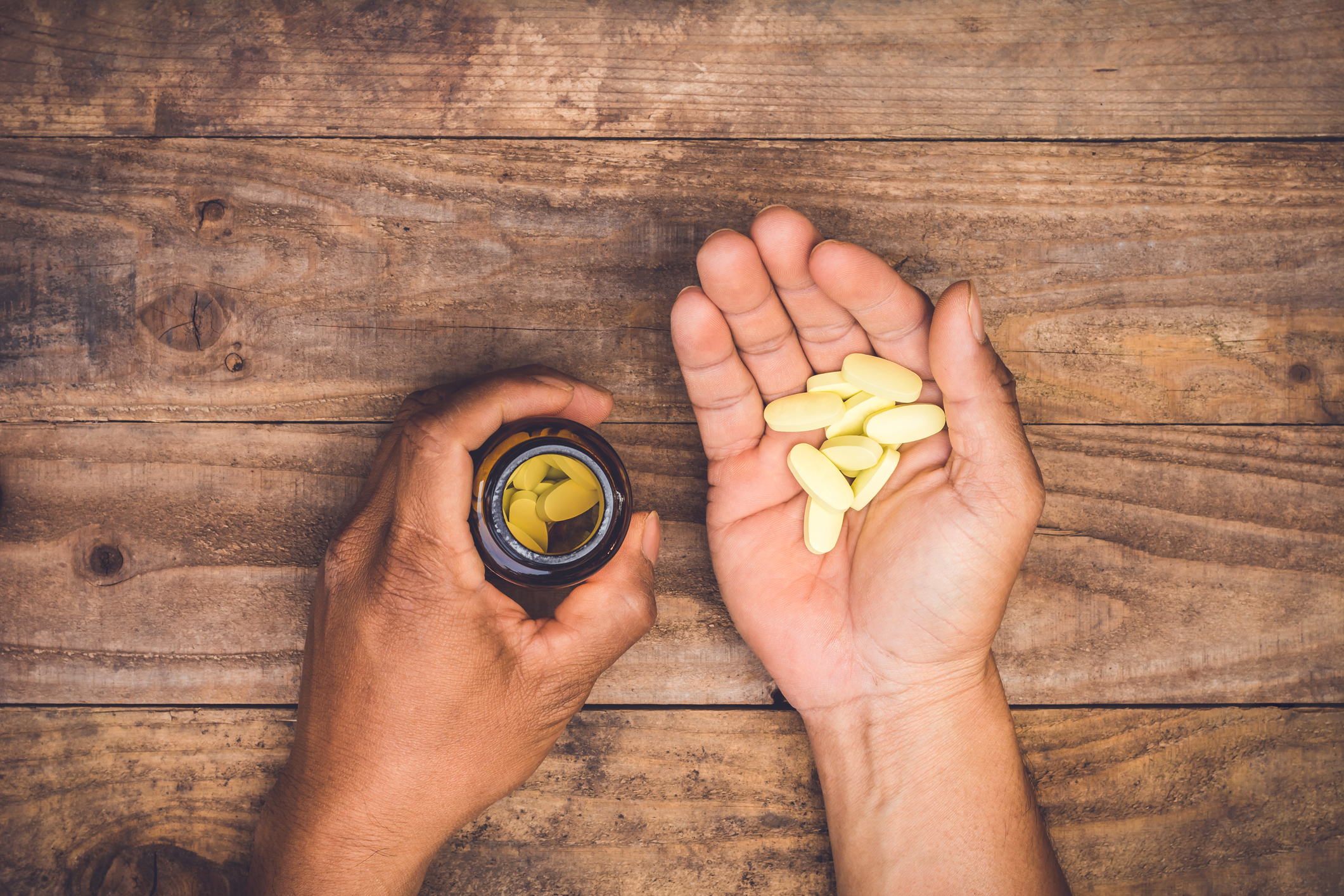 Hands holding some pills from a bottle Hands lying on a wooden table and holding some pills in one hand and an open pill bottle in the other.