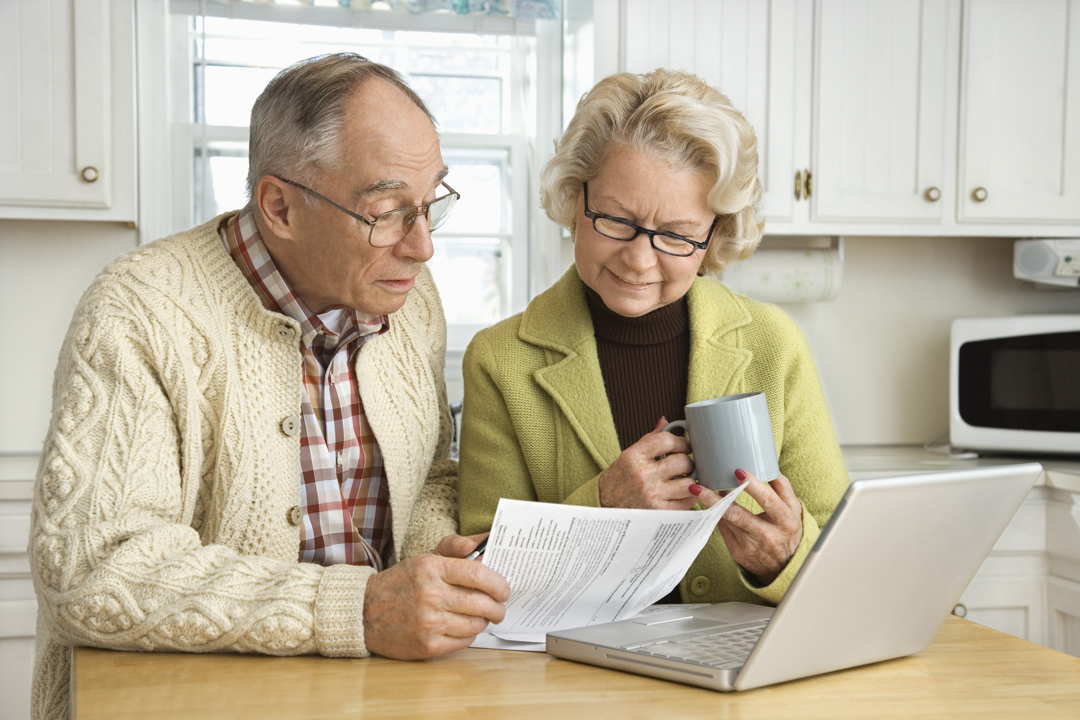 senior man and woman sitting at kitchen table and looking at papers in front of laptop Two people sitting at kitchen table and looking at papers in front of laptop.