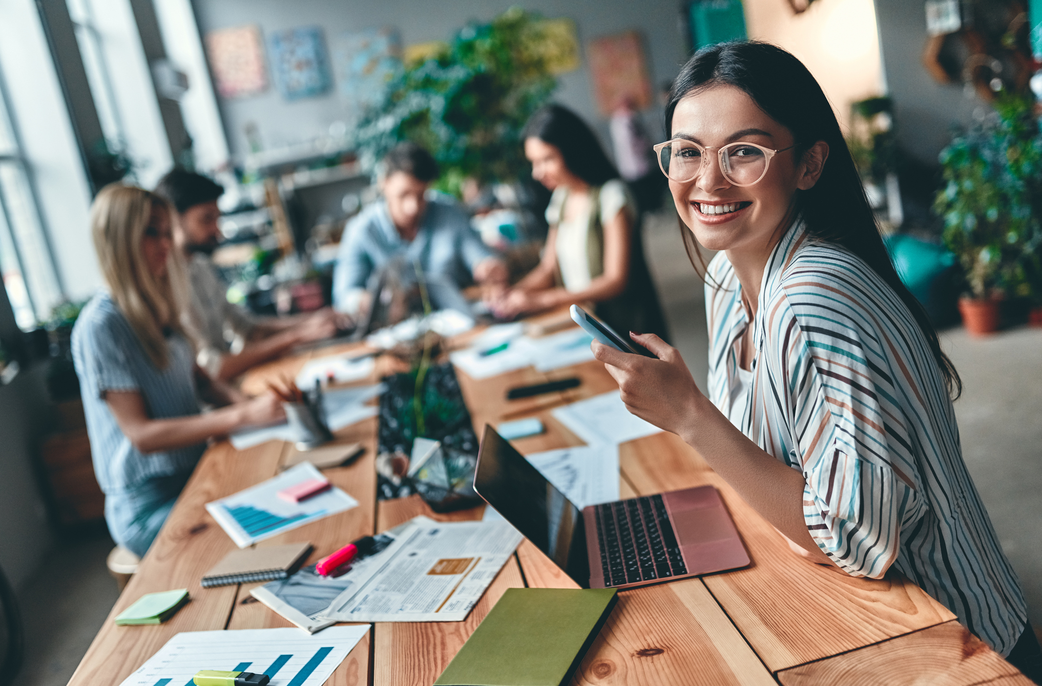 Woman in striped shirt and glasses smiling and sitting at long wooden table with other remote workers Person in striped shirt and glasses is smiling and sitting at long wooden table with other remote workers.