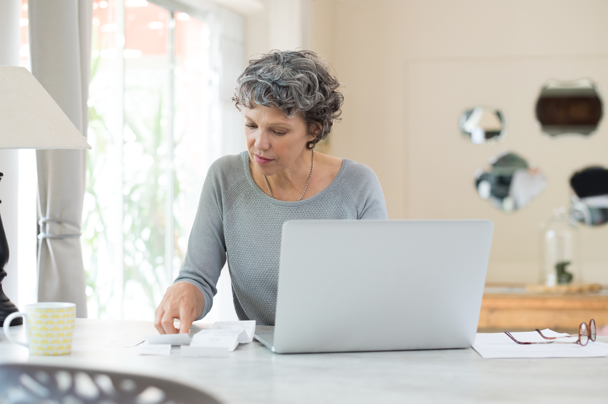 Older woman sitting at table in front of computer looking at documents.