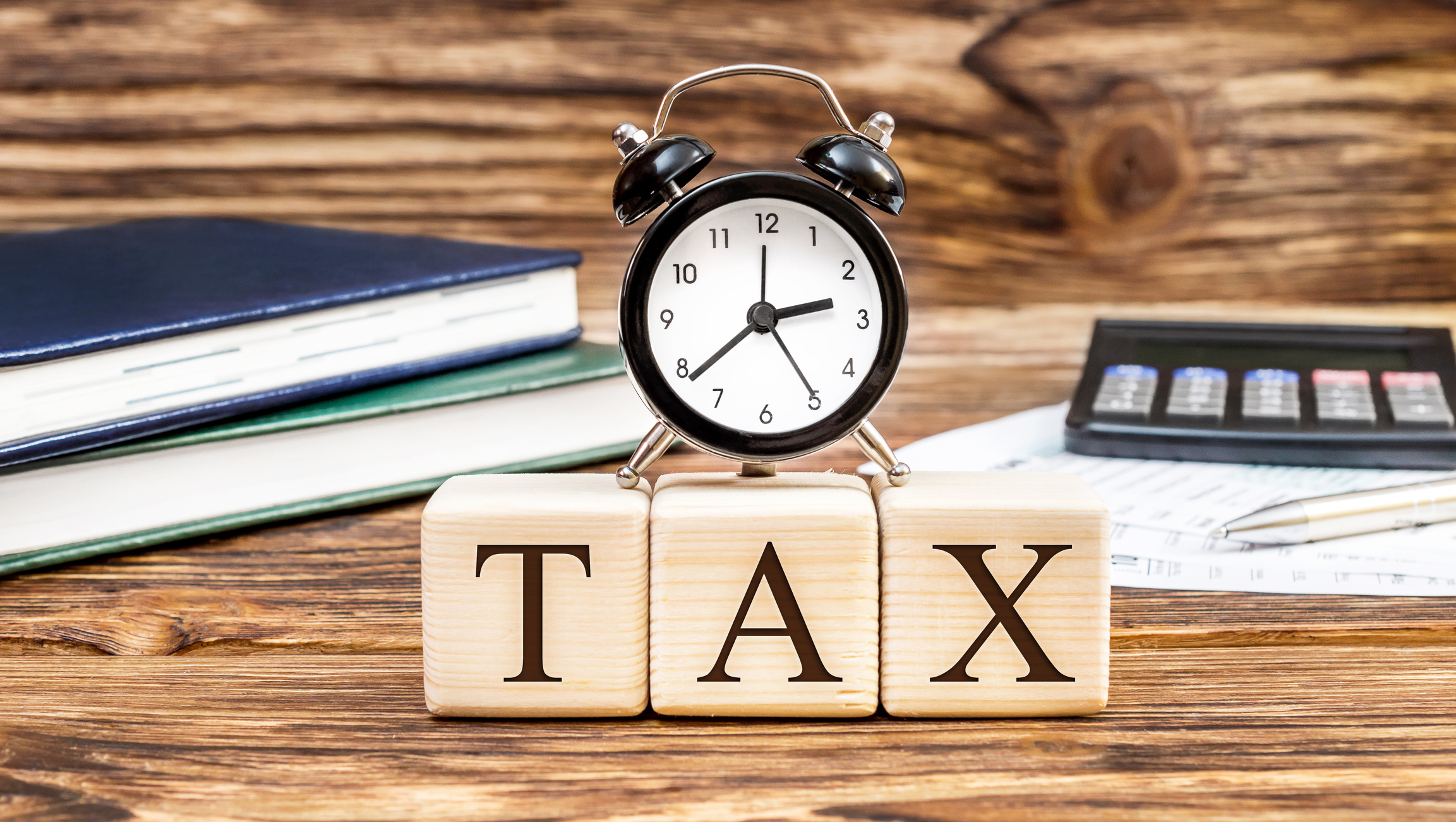 An alarm clock resting atop little wooden blocks that spell out TAX and sitting on a desk with books and a calculator. An alarm clock resting atop little wooden blocks that spell out TAX and sitting on a desk with books and a calculator.
