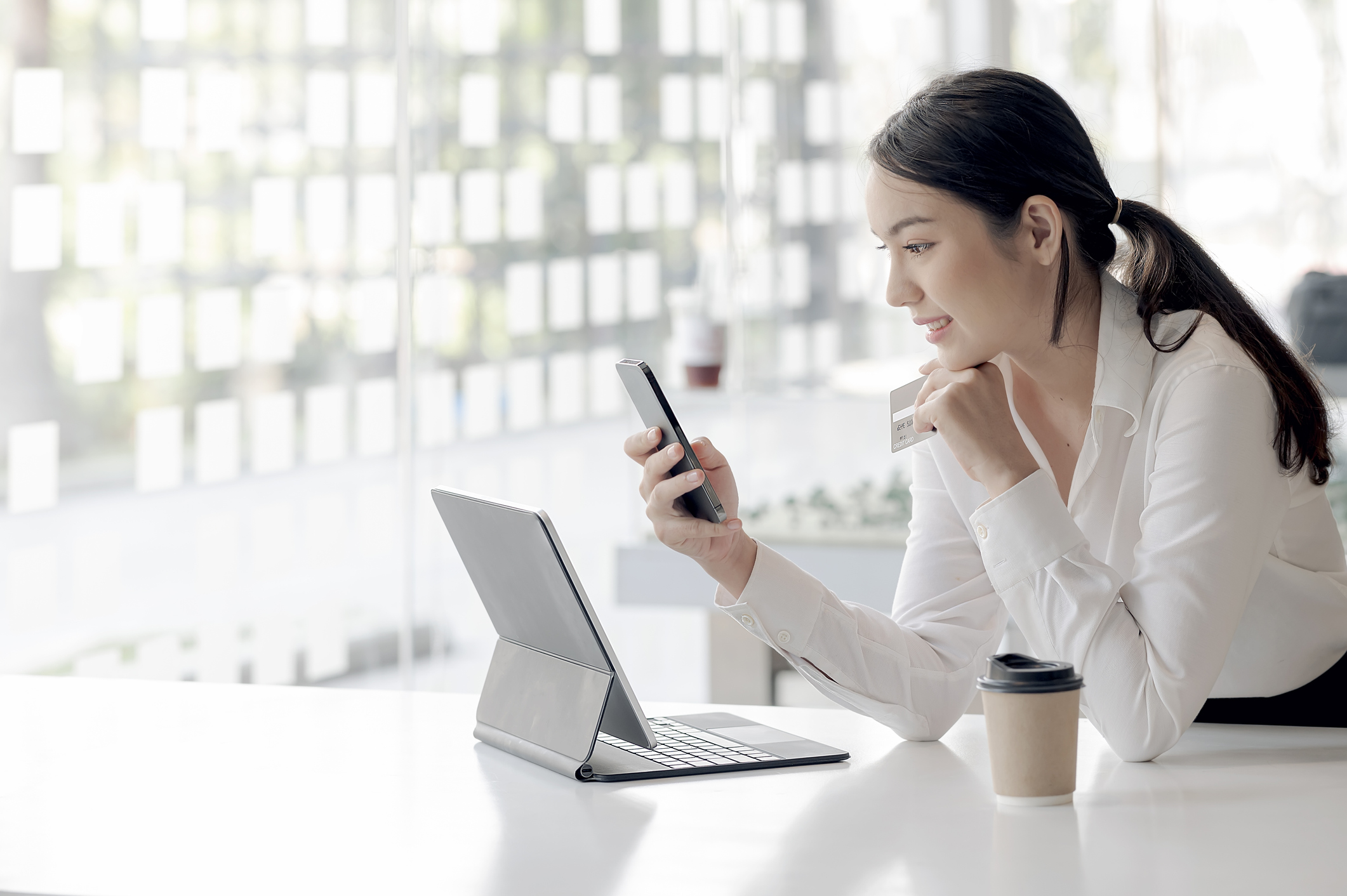 businesswoman_smiling_while_using_phone Person holding smartphone and credit card while sitting at office desk.