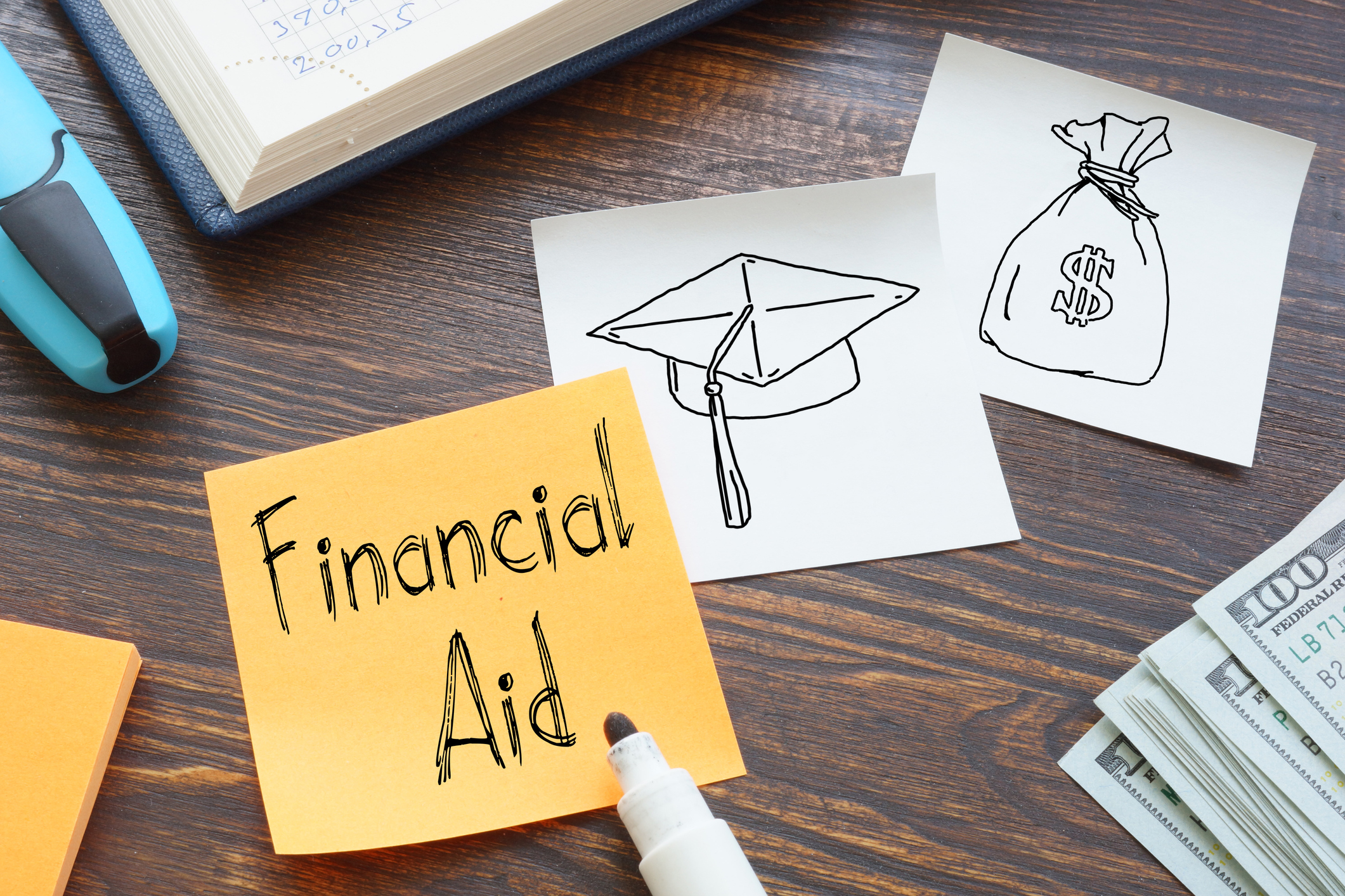 college university financial aid Financial aid written on a Post-It note lying on a desk next to a pile of hundred dollar bills and drawings of a cap and a bag of money.