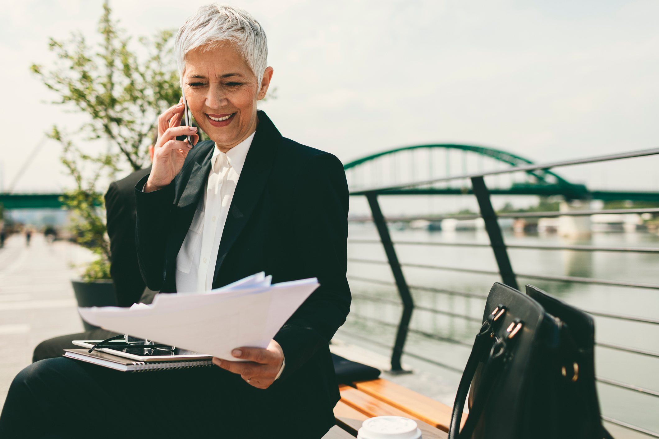 Mature senior businessperson on phone looking at documents Person sitting on a bench near a waterway and bridge while talking on the phone and looking at documents.