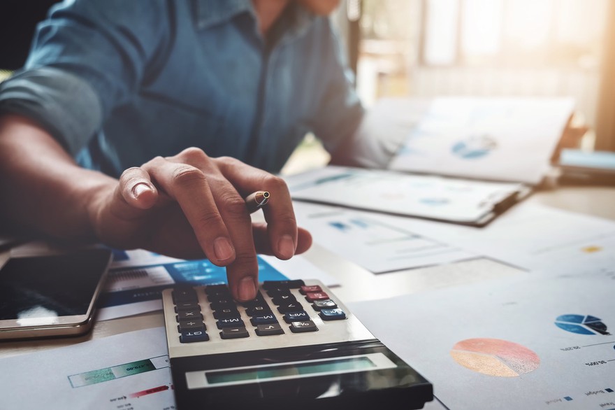 A person using a calculator with financial charts on the desk. A person using a calculator with financial charts on the desk.