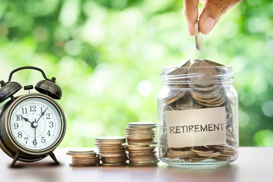 Hand placing a coin into a glass jar labeled "retirement" with stacked coins and an alarm clock next to it. Hand placing a coin into a glass jar labeled "retirement" with stacked coins and an alarm clock next to it.
