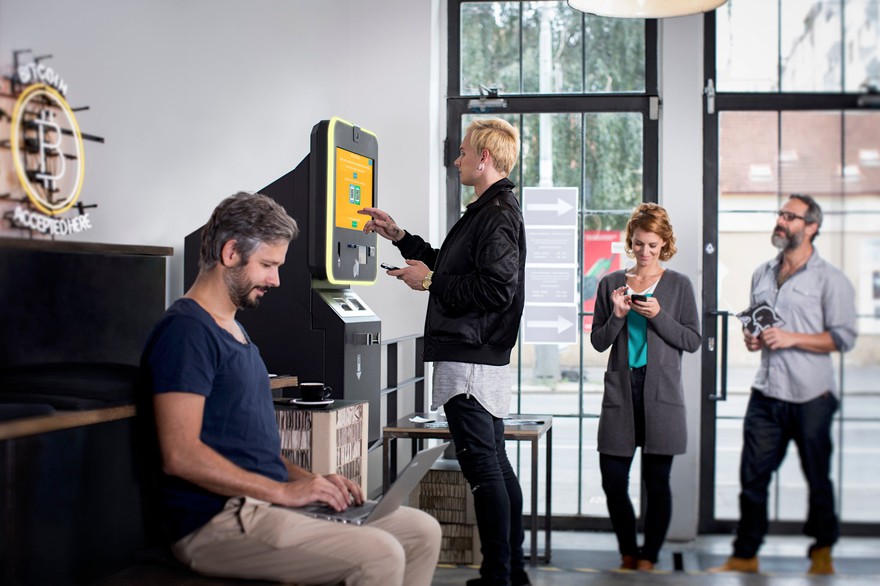 A man using a bitcoin ATM while two people wait in line. A man using a bitcoin ATM while two people wait in line.