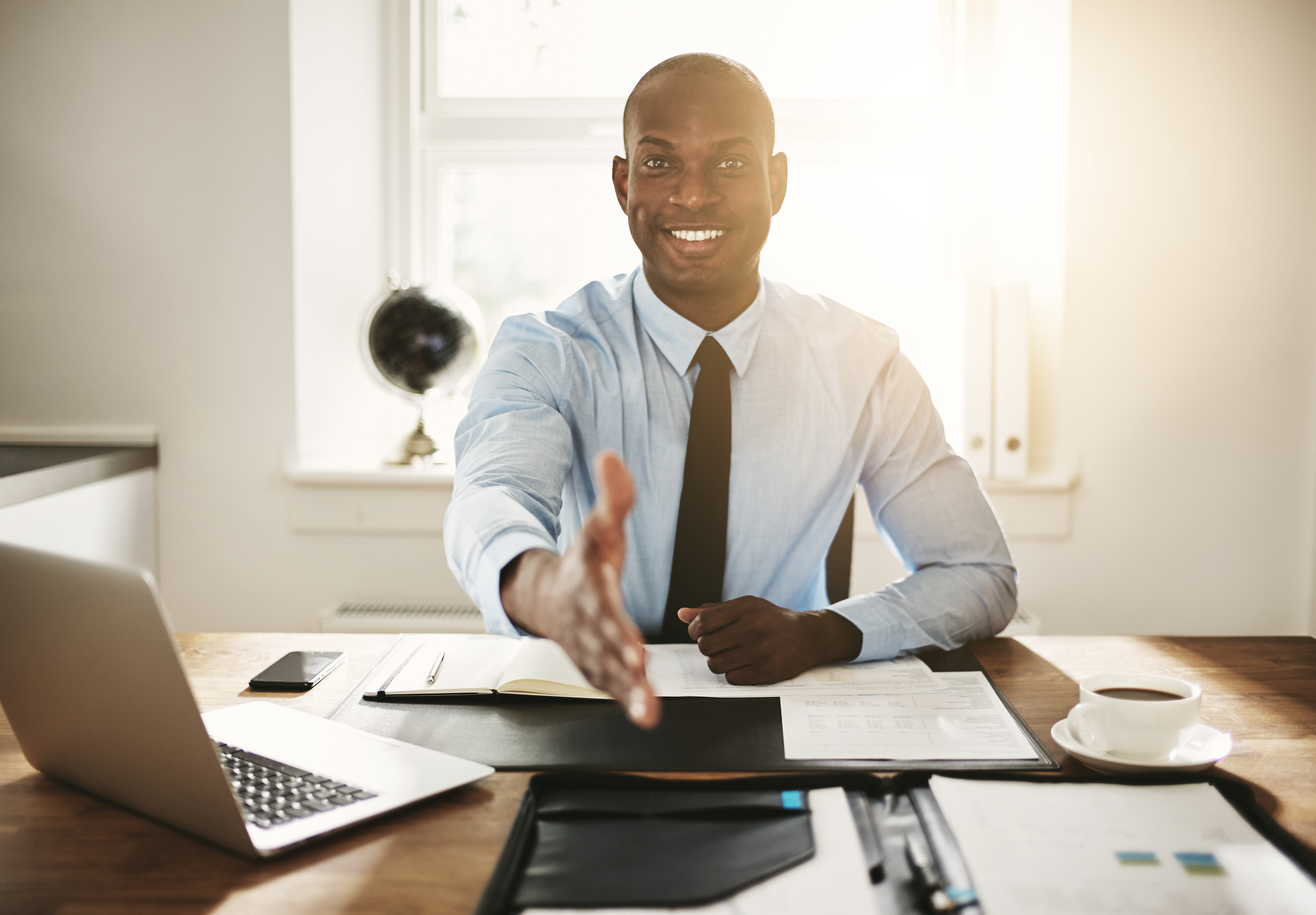 Professionally dressed man at desk extending a handshake to unseen individual Smiling business professional sitting at a desk and extending a handshake to unseen individual.