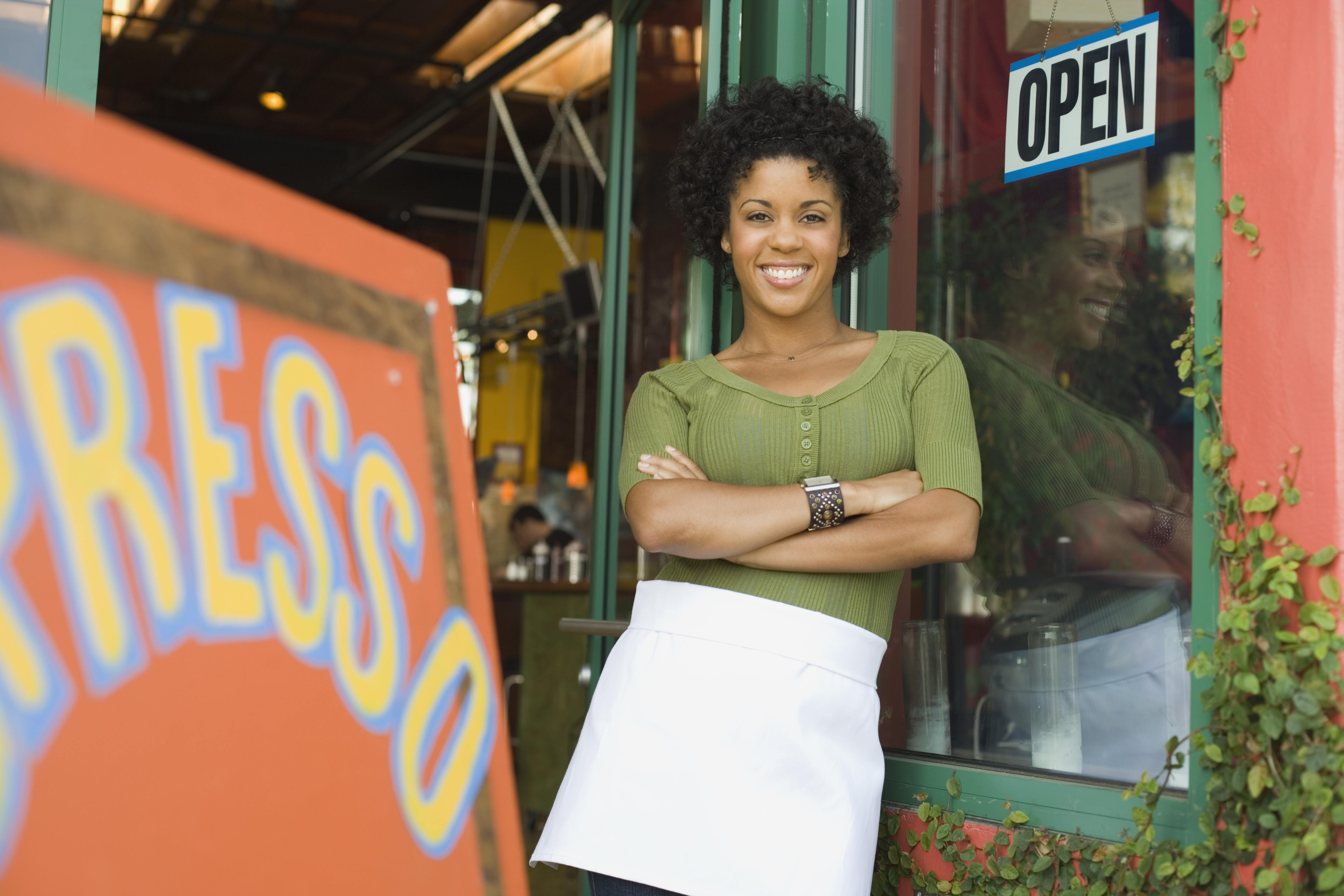 smiling woman wearing apron standing outside coffee shop with her arms folded -- entrepreneur small business POC Smiling person wearing apron and standing outside coffee shop has arms folded.