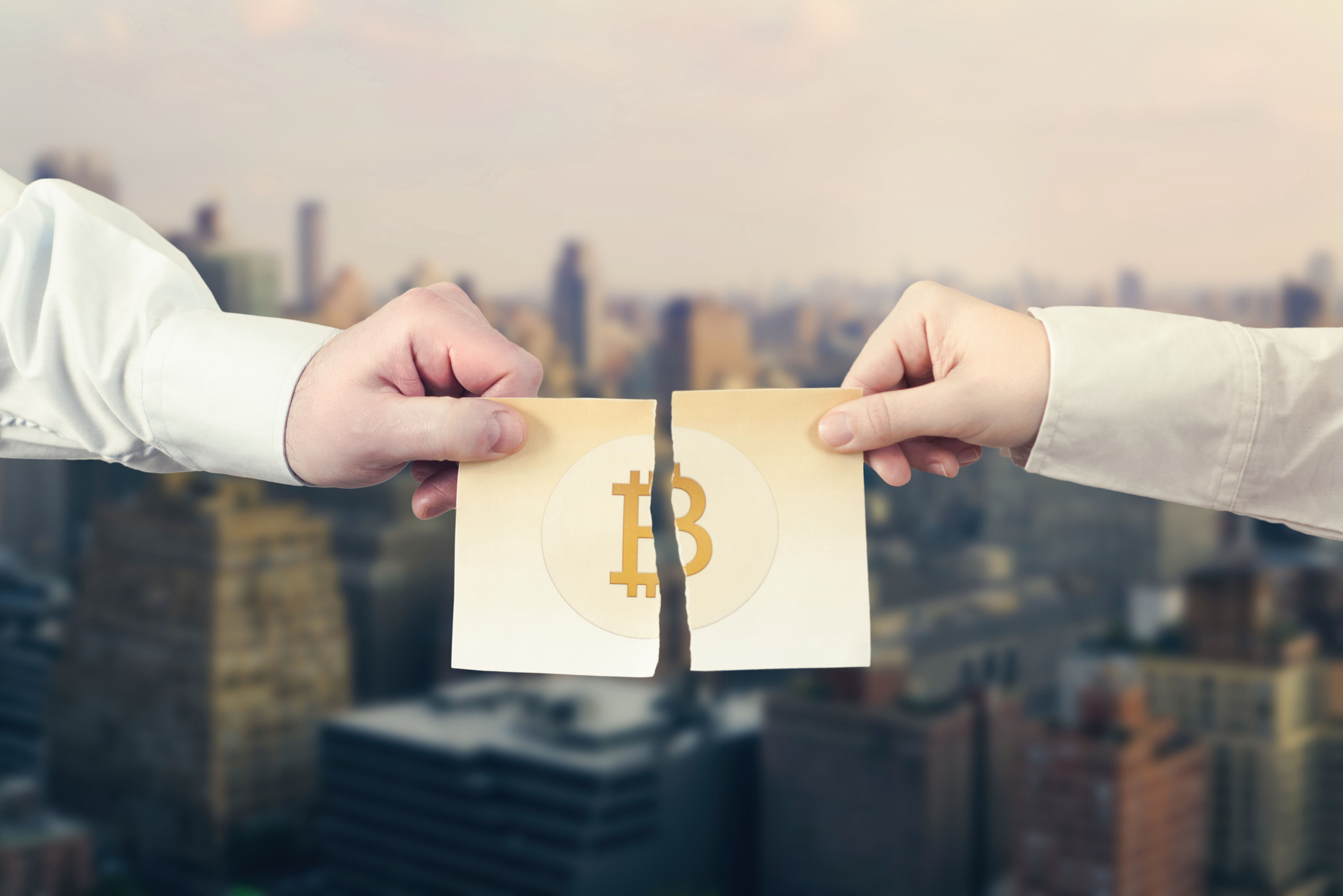 Splitting Bitcoin In Two Two hands pull on a paper with the Bitcoin logo, tearing it in half.