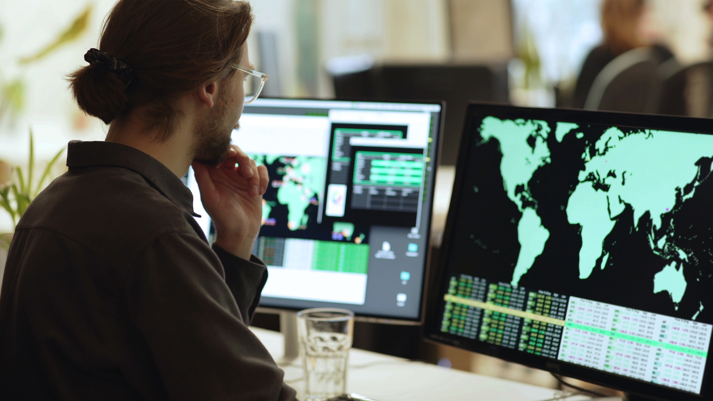 young_bearded_man_global_data_office_screens Person wearing glasses is surrounded by computer monitors in an office. In front of him there are screens showing maps of the world with associated data.